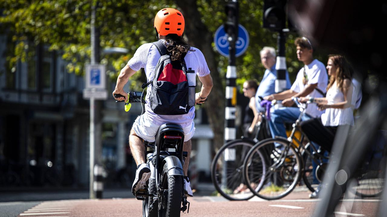 Ein Mann mit einem Elektrofahrrad im Zentrum von Amsterdam mit einem organgenen Helm. Im Hintergrund eine Gruppe von analogen Fahrradfahrerinnen.