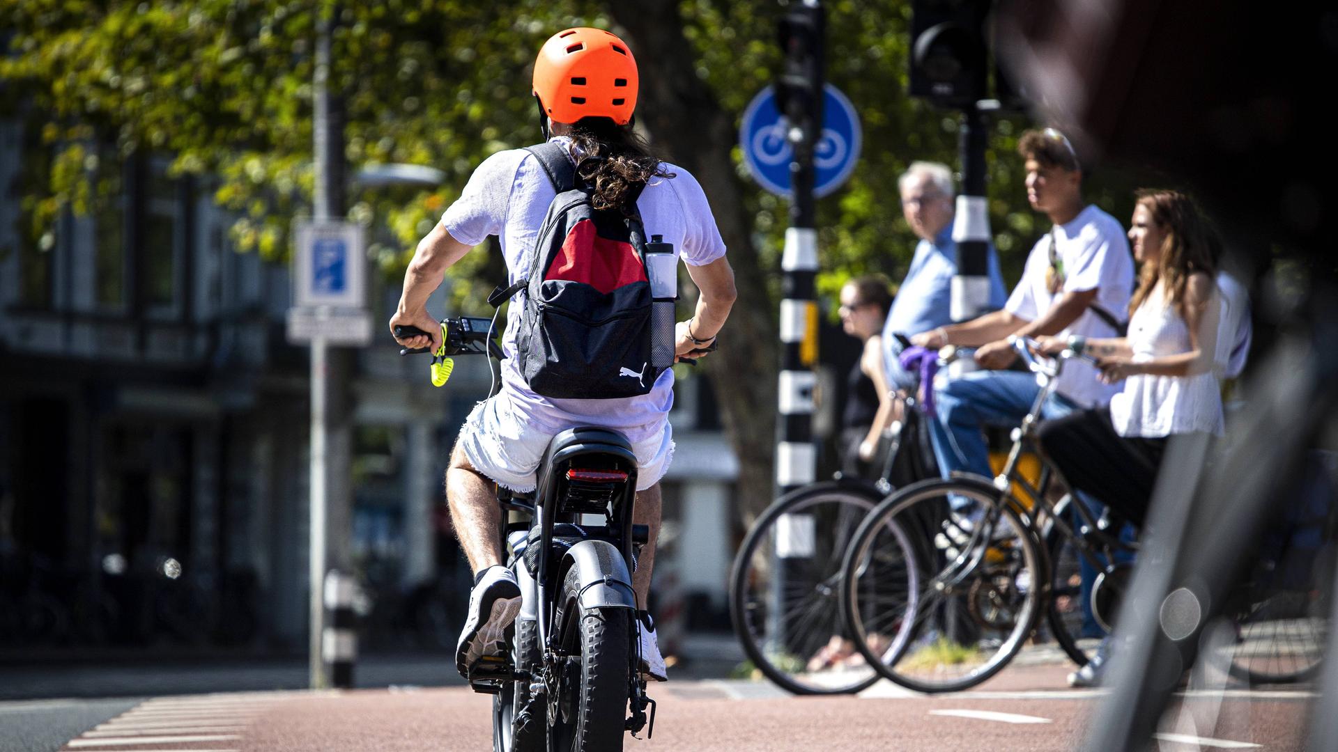 Ein Mann mit einem Elektrofahrrad im Zentrum von Amsterdam mit einem organgenen Helm. Im Hintergrund eine Gruppe von analogen Fahrradfahrerinnen.