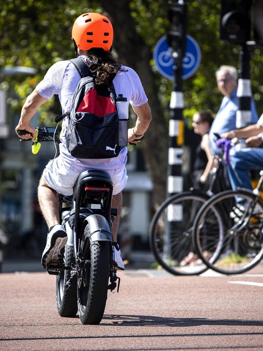 Ein Mann mit einem Elektrofahrrad im Zentrum von Amsterdam mit einem organgenen Helm. Im Hintergrund eine Gruppe von analogen Fahrradfahrerinnen. Ein Mann mit einem Elektrofahrrad im Zentrum von Amsterdam mit einem organgenen Helm. Im Hintergrund eine Gruppe von analogen Fahrradfahrerinnen.