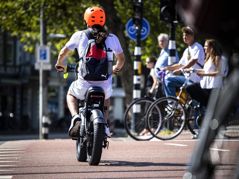 Ein Mann mit einem Elektrofahrrad im Zentrum von Amsterdam mit einem organgenen Helm. Im Hintergrund eine Gruppe von analogen Fahrradfahrerinnen. Ein Mann mit einem Elektrofahrrad im Zentrum von Amsterdam mit einem organgenen Helm. Im Hintergrund eine Gruppe von analogen Fahrradfahrerinnen.