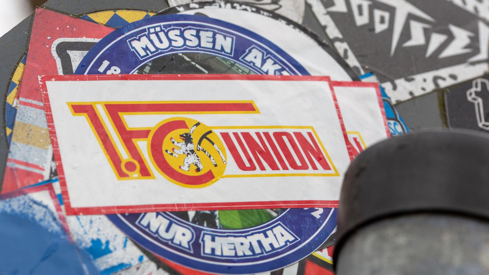 Aufkleber mit Motiven von Hertha BSC und dem 1. FC Union Berlin sind auf einem Straßenverkehrschild übereinander angeklebt. 