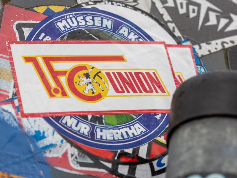 Aufkleber mit Motiven von Hertha BSC und dem 1. FC Union Berlin sind auf einem Straßenverkehrschild übereinander angeklebt. 