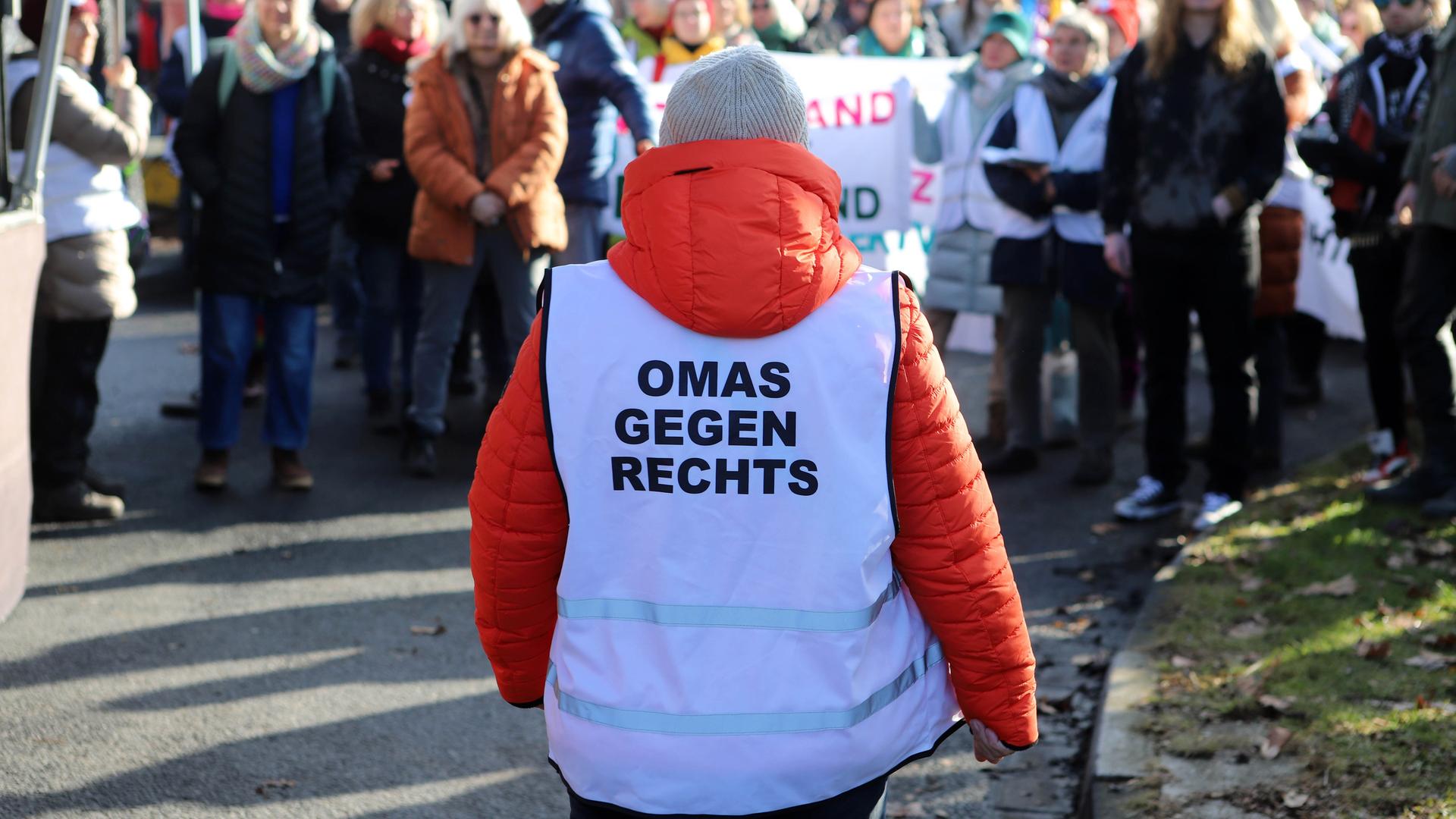 Eine Gruppe steht auf der Straße. Es ist eine Demonstration. Vor ihnen steht eine Frau mit orangener Jacke und einer Weste mit dem Aufdruck "Omas gegen rechts". 