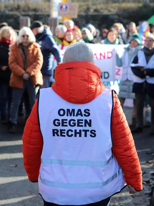 Eine Gruppe steht auf der Straße. Es ist eine Demonstration. Vor ihnen steht eine Frau mit orangener Jacke und einer Weste mit dem Aufdruck "Omas gegen rechts". 