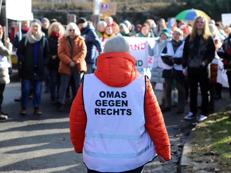 Eine Gruppe steht auf der Straße. Es ist eine Demonstration. Vor ihnen steht eine Frau mit orangener Jacke und einer Weste mit dem Aufdruck "Omas gegen rechts". 