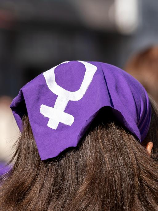 Das Foto zeigt den Hinterkopf einer weiblichen Person mit langem, dunklem Haar. Die Person trägt ein lilafarbenes Kopftuch, auf das in weißer Farbe das Gender-Symbol für Weiblichkeit gemalt wurde. 