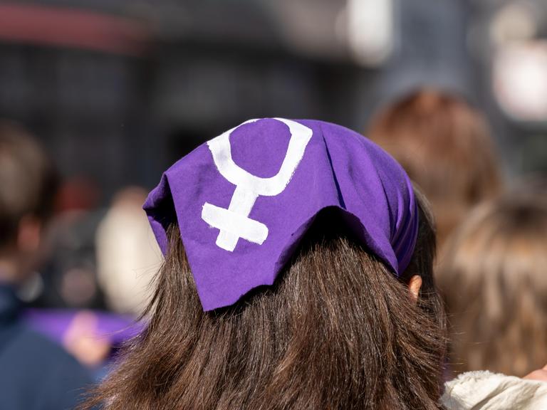 Das Foto zeigt den Hinterkopf einer weiblichen Person mit langem, dunklem Haar. Die Person trägt ein lilafarbenes Kopftuch, auf das in weißer Farbe das Gender-Symbol für Weiblichkeit gemalt wurde. 