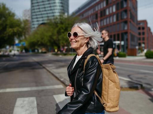 Frau mit schicker Sonnenbrille im mittleren Alter überquert eine Straße in einer Stadt, sie wirkt dynamisch und lächelt. (Symbolbild)