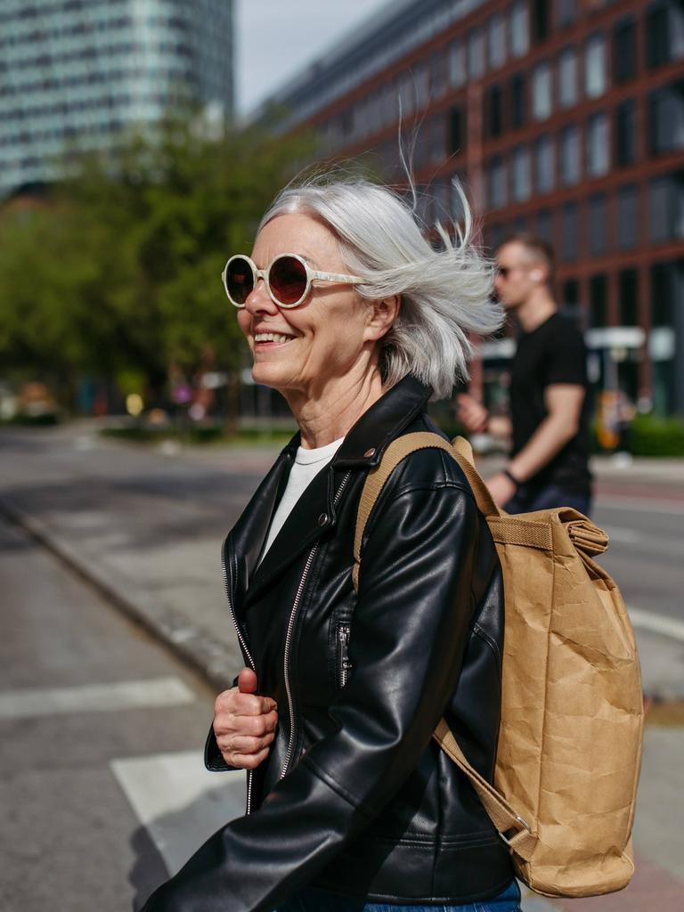 Frau mit schicker Sonnenbrille im mittleren Alter überquert eine Straße in einer Stadt, sie wirkt dynamisch und lächelt. (Symbolbild)