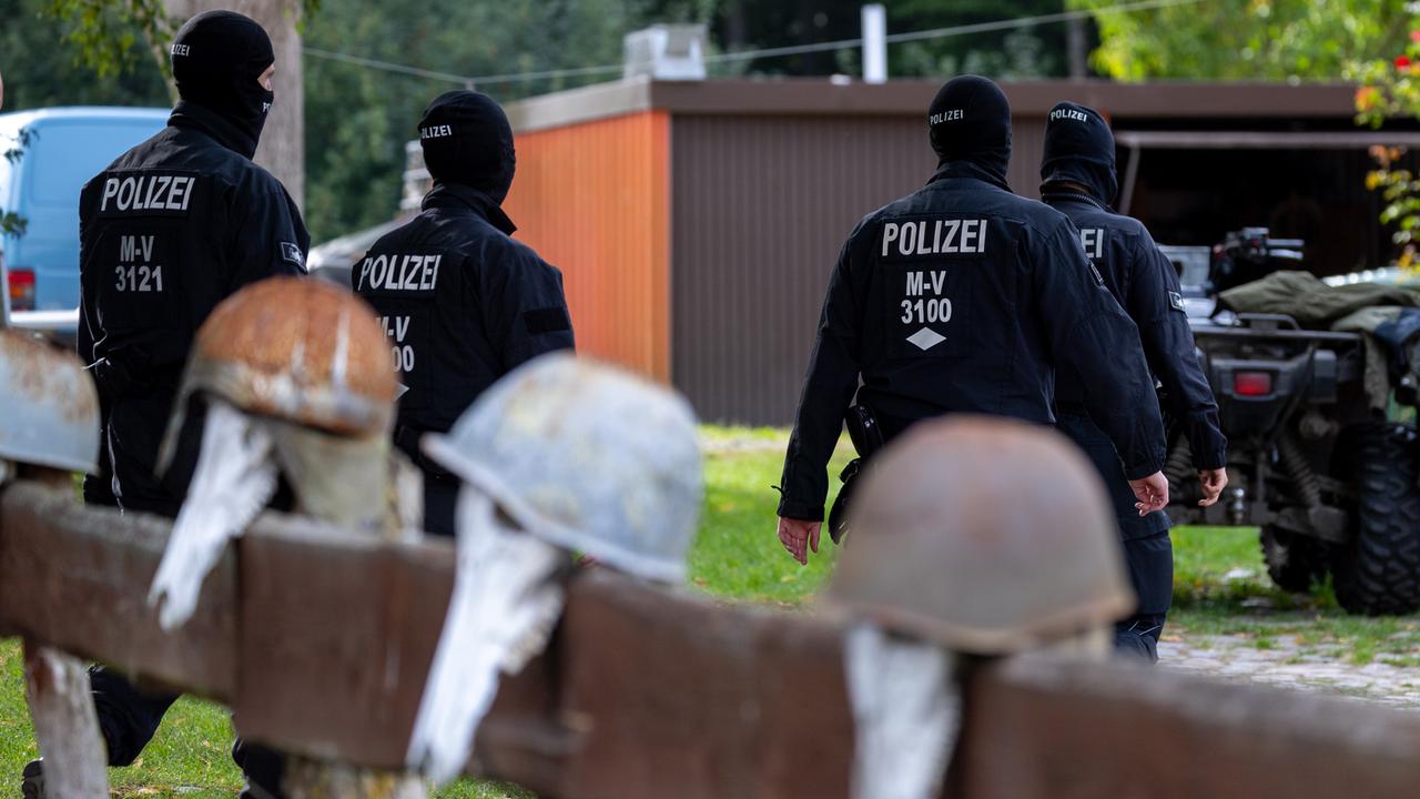 Vier Polizisten mit dunkler Kleidung und Gesichtshauben sind bei einer Durchsuchung auf einem Grundstück zu sehen. Im Vordergrund hängen vier verrostete Stahlhelme auf einem Zaun. 