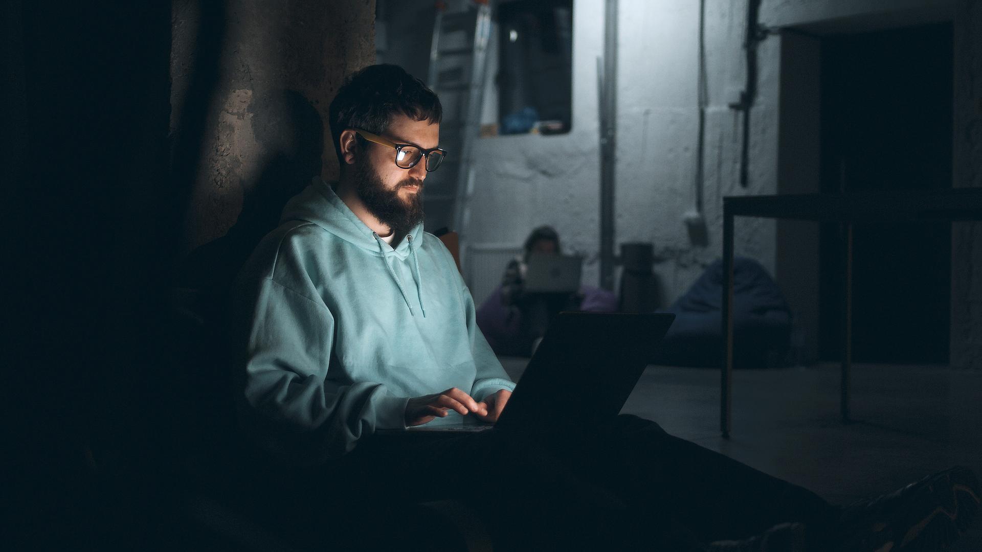 Ein junger Mann mit Bart sitzt in einem dunklen Bunker in der Ukraine an seinem Laptop.