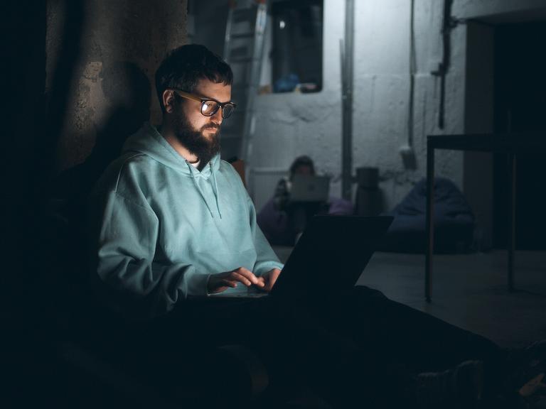 Ein junger Mann mit Bart sitzt in einem dunklen Bunker in der Ukraine an seinem Laptop.
