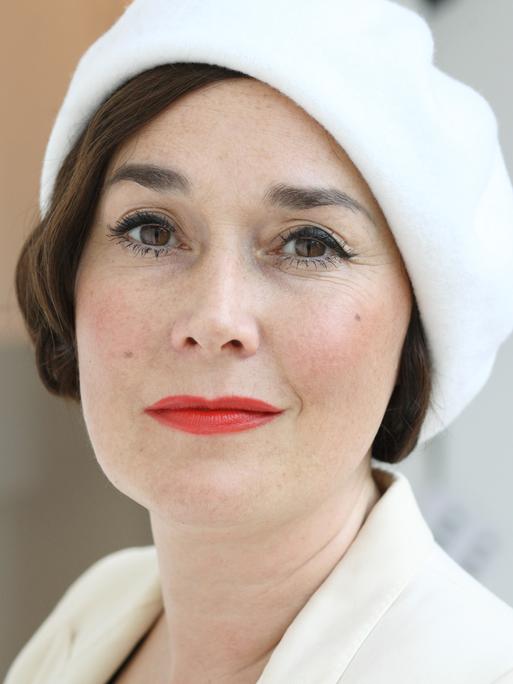 Porträtfoto einer weißen Frau mit kurzen, braunen Haaren, braunen Augen und rot geschminkten Lippen. Sie trägt eine elegante weiße Mütze, die leicht schräg auf ihrem Kopf sitzt. Es handelt sich um Fiona Bennett.