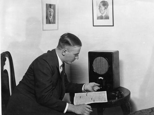 Historisches Foto eines Mannes in schwarzem Anzug, der an einem Wohnzimmertisch vor einem Volksempfänger sitzt.