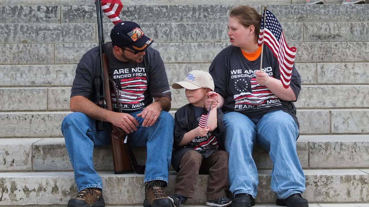 Eine junge Familie mit amerikanischer Flaggen sitzt auf Stufen vor einem Gebäude. Der Vater lehnt an einem Gewehr, sie unterhalten sich.