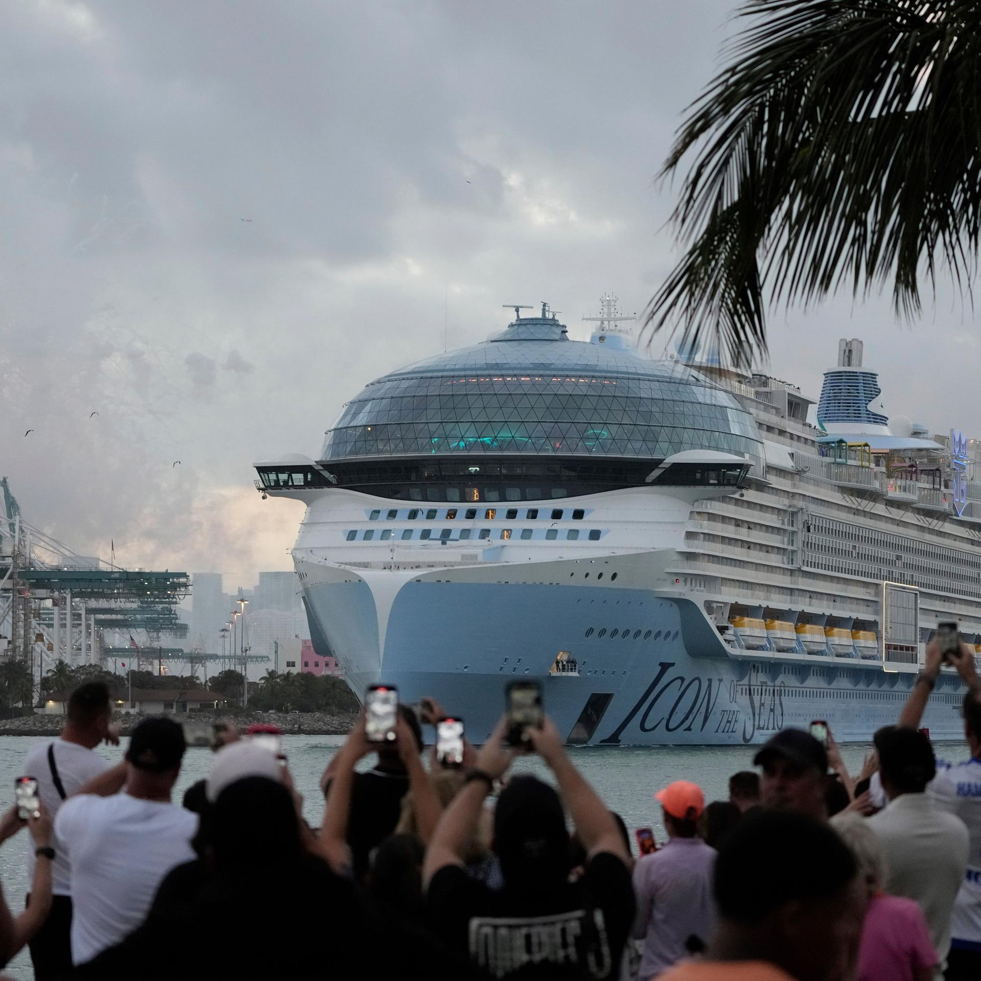 Menschen stehen in Miami am Ufer und fotografieren und filmen im Januar 2024 mit ihren Smartphones die Jungfernfahrt des weltgrößten Kreuzfahrtschiffes "Icon of the Seas",
