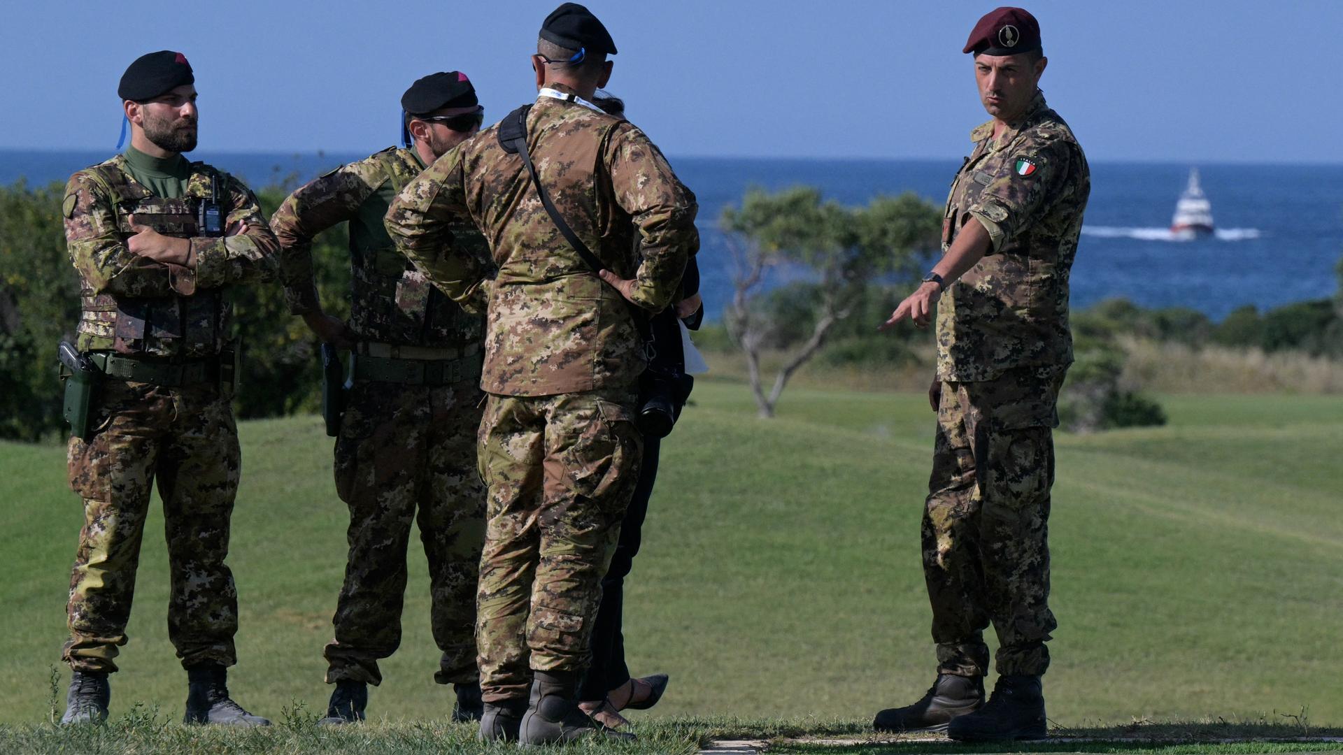 Italienische Soldaten stehen auf einer Wiese und diskutieren. Im Hintergrund ist ein Boot der Küstenwache zu sehen.