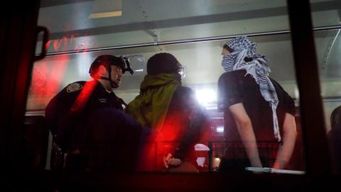 Das Bild zeigt einen Demonstranten mit Palästinensertuch, der mit mit Kabelbindern an den Händen gefesselt von US-Polizeibeamten in einem Bus geleitet wird.
