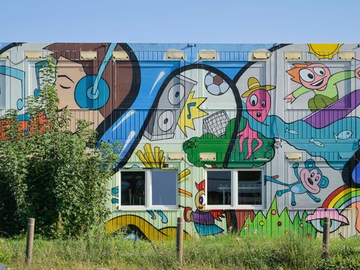 Ein mit bunten Comicmotiven - darunter ein Surfer, ein Teddy, ein Junge mit Kopfhörern und eine fußballspielenden Krake - bemalter Container einer Grund- und Oberschule in Schwanebeck in Brandenburg.