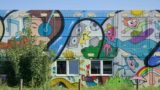 Ein mit bunten Comicmotiven - darunter ein Surfer, ein Teddy, ein Junge mit Kopfhörern und eine fußballspielenden Krake - bemalter Container einer Grund- und Oberschule in Schwanebeck in Brandenburg.