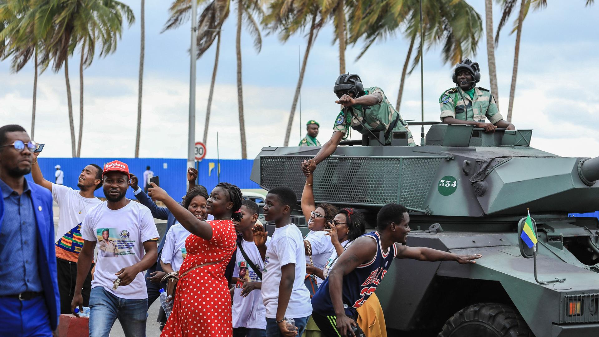 Menschen auf der Straße posieren mit Soldaten, die sich aus einem Panzer zu ihnen herunter beugen.