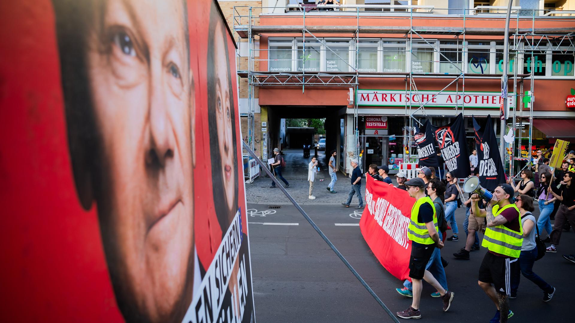 Der Zug einer Demonstration linker Gruppen unter dem Motto "Für Frieden und soziale Gerechtigkeit" zieht in Berlin-Wedding an einem Wahlkampfplakat der SPD mit Bundeskanzler Scholz und der Spitzenkandidatin für die Europawahl, Barley, vorbei. 