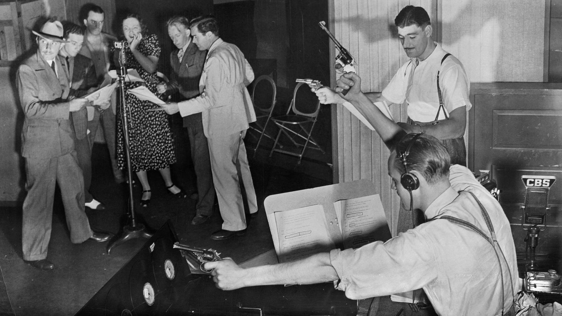Schwarzweißfoto eines Hörspielstudios. Schauspieler stehen um ein Mikrofon herum, während zwei Tonassistenten Pistolen abfeuern.