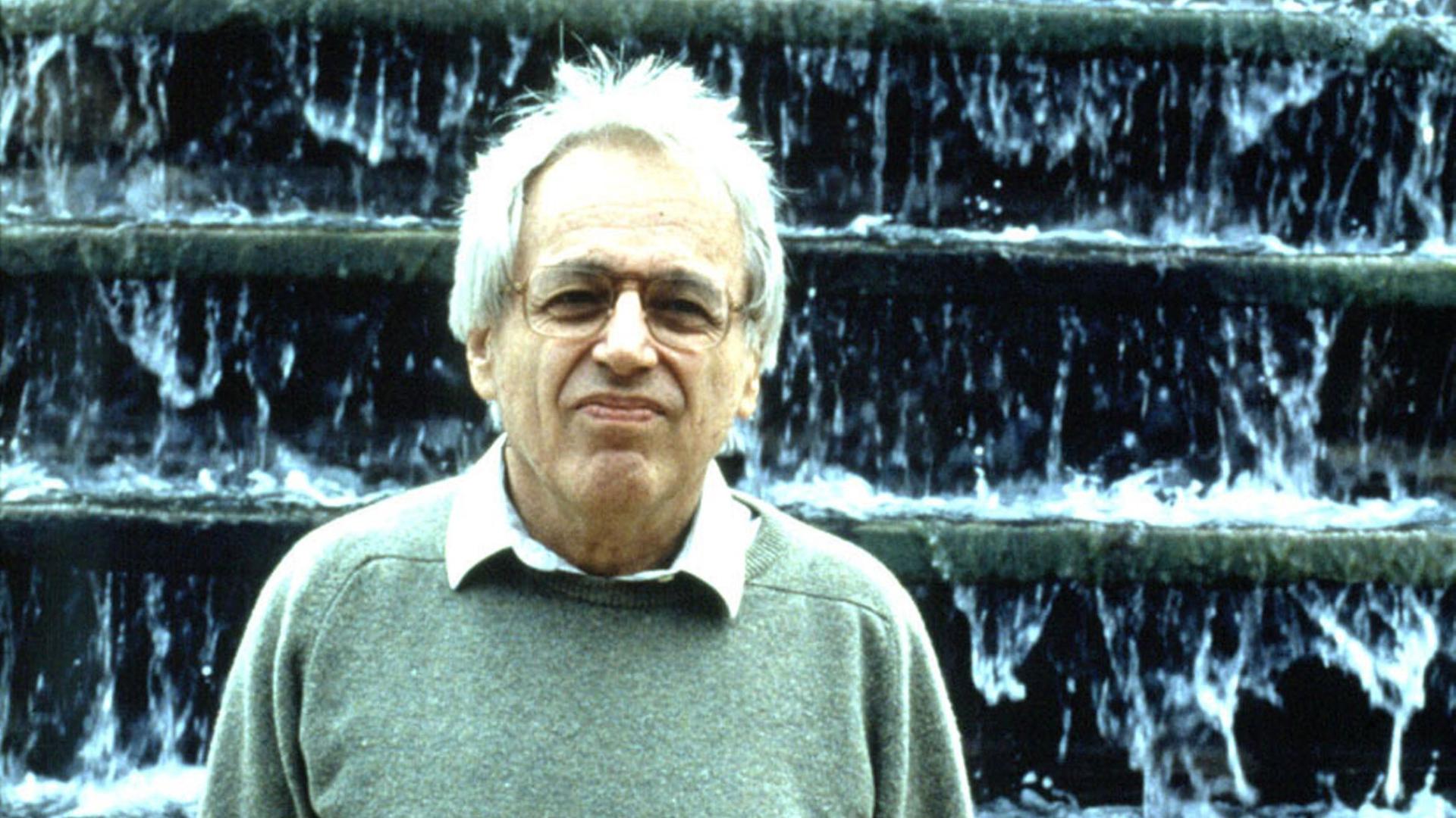 György Ligeti steht mit einem grünlichen Pullover und weißem Haar vor einer Wasserkunst, die wie ein großer Wasserfall aussieht. 