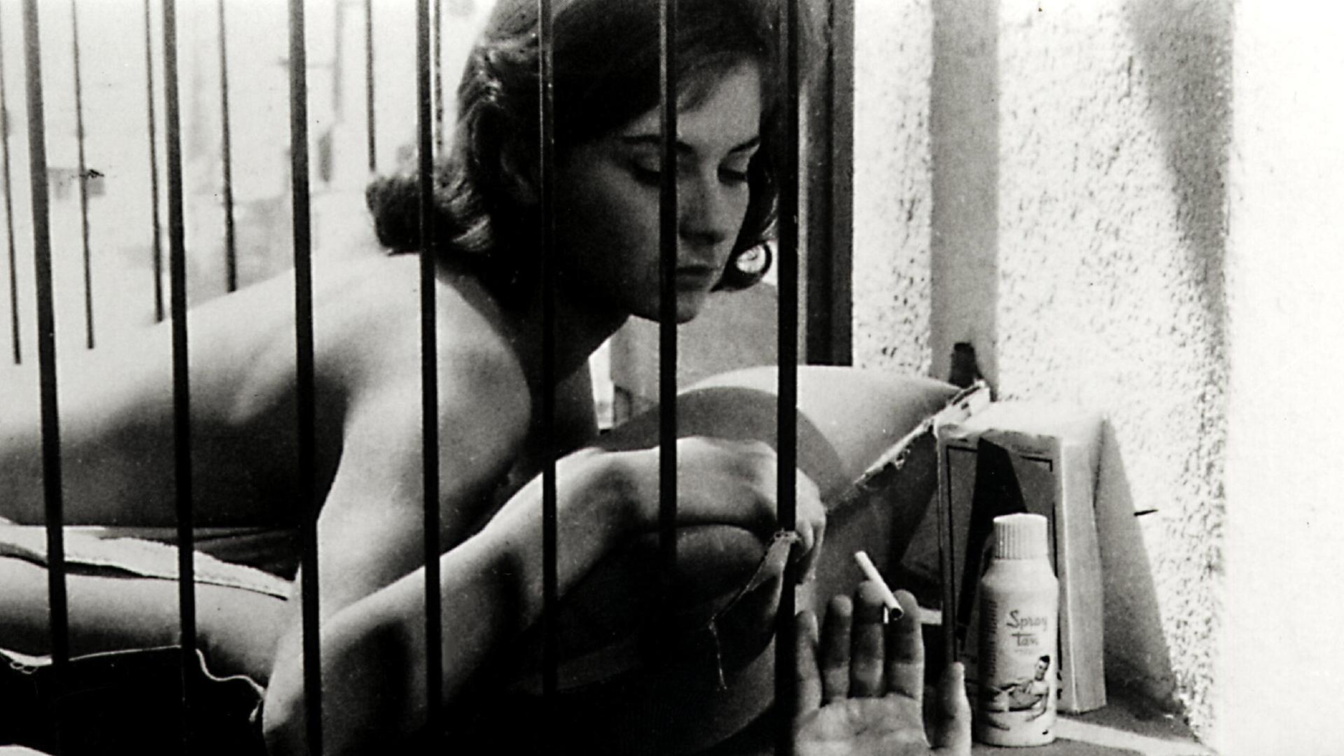 Juliette Mayniel liegt mit nacktem Oberkörper auf einer Matratze hinter Gittern. Jemand reicht ihr eine Zigarette.