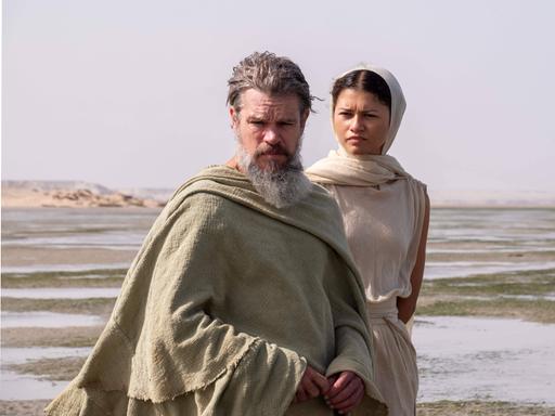 Matt Damon als Odysseus und Zendaya als Athena stehen vor einer Wüsten-ähnlichen Landschaft. Matt Damon als Odysseus und Zendaya als Athena stehen vor einer Wüsten-ähnlichen Landschaft.