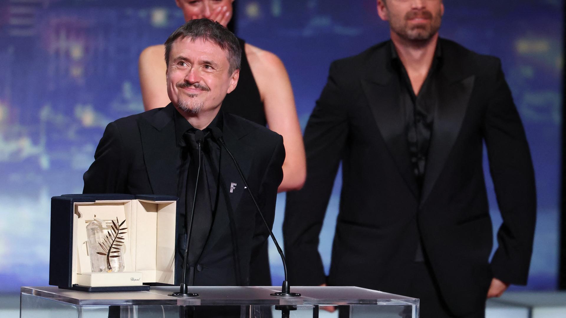 Cristian Mungiu steht an einem Rednerpult und lächelt. Vor ihm steht die Goldene Palme in einer Schatulle. 
