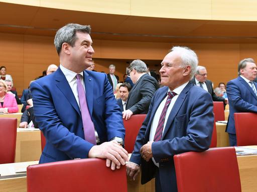 Bayerns Ministerpräsident Markus Söder steht neben dem ehemaligen CSU-Vorsitzenden Erwin Huber in einem Parlamentsaal. Söder legt die Hände auf einen Stuhl und blickt in die Ferne, Huber an ihm vorbei.