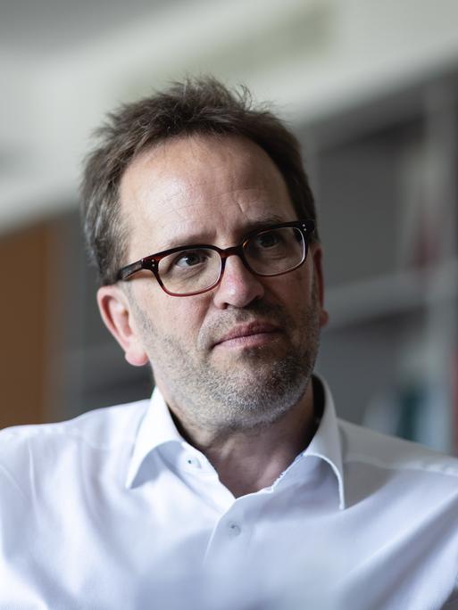 Klaus Müller sitzt in einem Raum, vermutlich auf einem Stuhl. Rechts von ihm ist ein Anschnitt eines Regals verschwommen zu sehen. Er trägt ein weißes Hemd und eine braune Brille und schaut nachdenklich zur rechten Seite aus dem Bild.