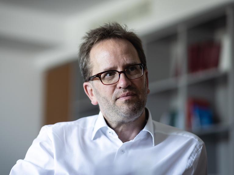 Klaus Müller sitzt in einem Raum, vermutlich auf einem Stuhl. Rechts von ihm ist ein Anschnitt eines Regals verschwommen zu sehen. Er trägt ein weißes Hemd und eine braune Brille und schaut nachdenklich zur rechten Seite aus dem Bild.