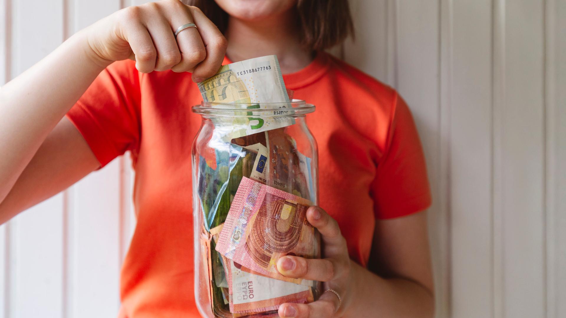 Eine lachende Frau mit einem orange-roten T-Shirt nimmt einen fünf Euroschein aus einem Glas, in dem auch noch andere Euroscheine sind.