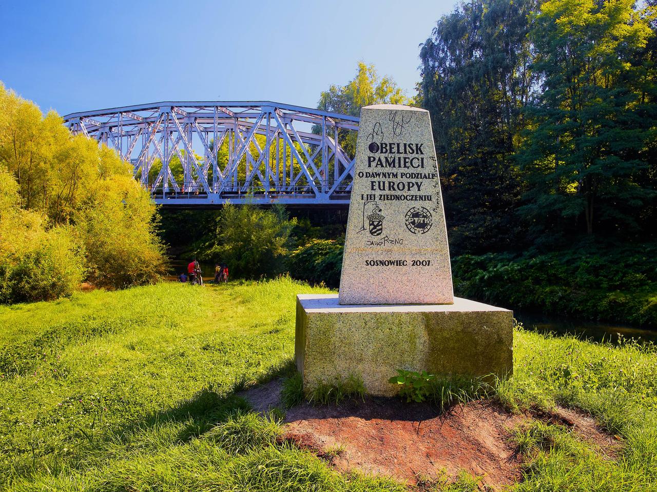 Das sogenannte „Dreikaisereck“ in Polen. Auf einer Wiese steht in Gedenkstein, im Hintergrund ist eine Brücke zu sehen. 