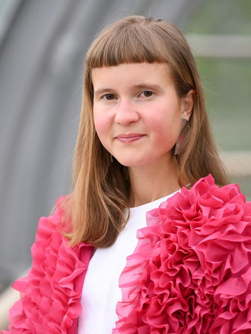 Eine junge Frau mit ungewöhnlicher Pnyfrisur und einer pinken Jacke schaut in die Kamera: die Schriftstellerin Caroline Wahl