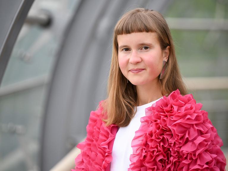 Eine junge Frau mit ungewöhnlicher Pnyfrisur und einer pinken Jacke schaut in die Kamera: die Schriftstellerin Caroline Wahl