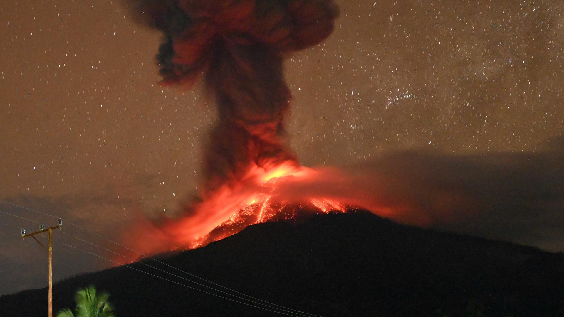 Das Bild zeigt den Kegel des Vulkans Lewotobi Laki-laki in der Nacht. Bei dem Ausbruch steigt eine hohe Aschewolke in den Himmel, und glühende Lava rinnt die Hänge hinunter. Das Bild zeigt den Kegel des Vulkans Lewotobi Laki-laki in der Nacht. Bei dem Ausbruch steigt eine hohe Aschewolke in den Himmel, und glühende Lava rinnt die Hänge hinunter.