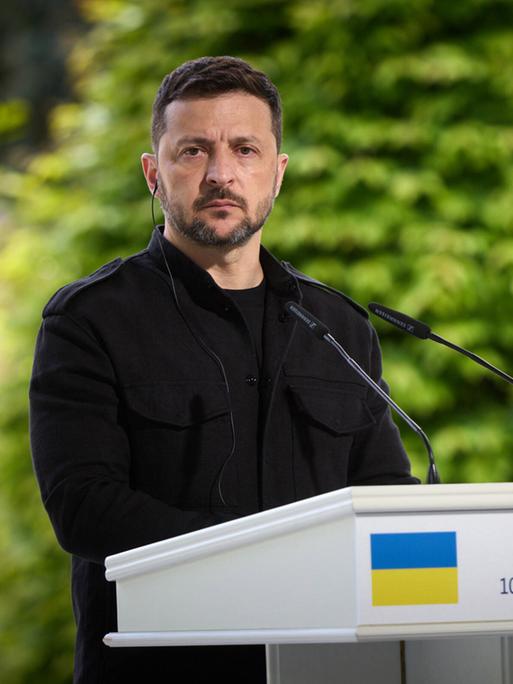 Ukraines Präsident Wolodymyr Selenskyj steht an einem Pult mit Mikrofon und blickt ernst in die Kamera