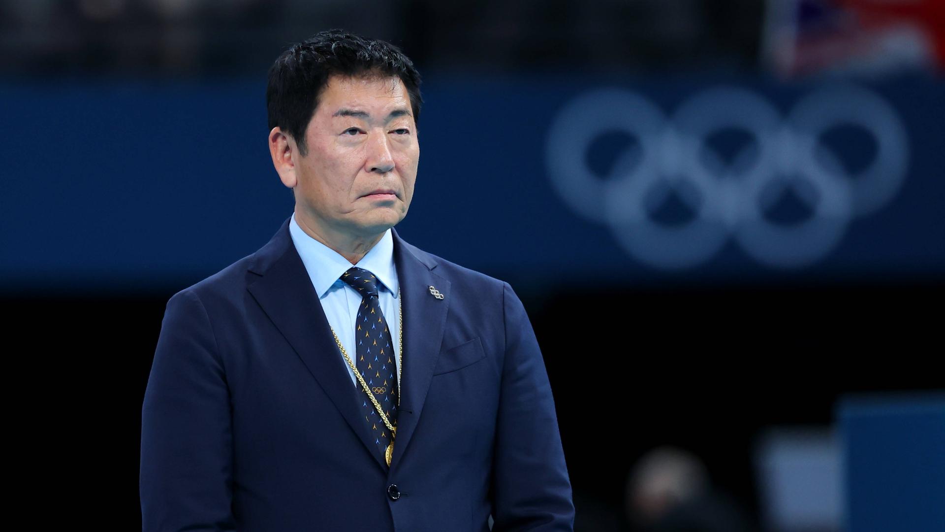 Morinari Watanabe steht im dunkelblauen Anzug vor einem Banner mit den olympischen Ringen.