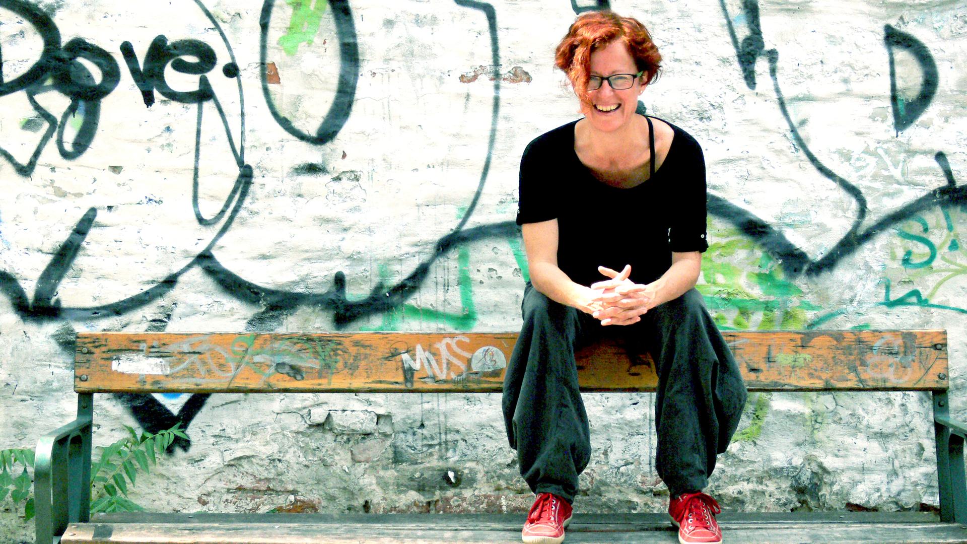 Eine Frau mit kurzen roten Haaren und Brille sitzt vor einer bemalten Wand lachend auf der Rücklehne einer Parkbank
