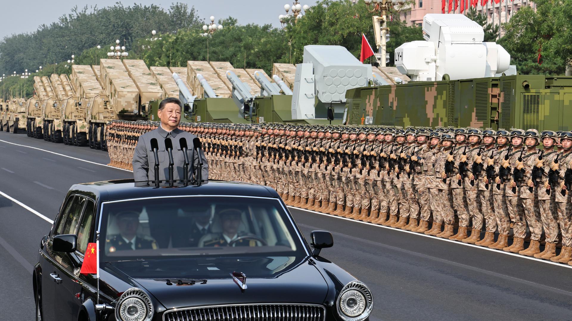 Der chinesische Präsident Xi steht in einem fahrenden Auto mit Mikrofonen. Er fährt an sehr vielen Soldaten und großen Waffen vorbei. Der chinesische Präsident Xi steht in einem fahrenden Auto mit Mikrofonen. Er fährt an sehr vielen Soldaten und großen Waffen vorbei.