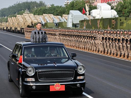 Der chinesische Präsident Xi steht in einem fahrenden Auto mit Mikrofonen. Er fährt an sehr vielen Soldaten und großen Waffen vorbei.