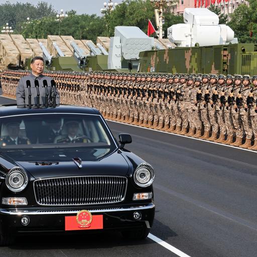 Der chinesische Präsident Xi steht in einem fahrenden Auto mit Mikrofonen. Er fährt an sehr vielen Soldaten und großen Waffen vorbei.