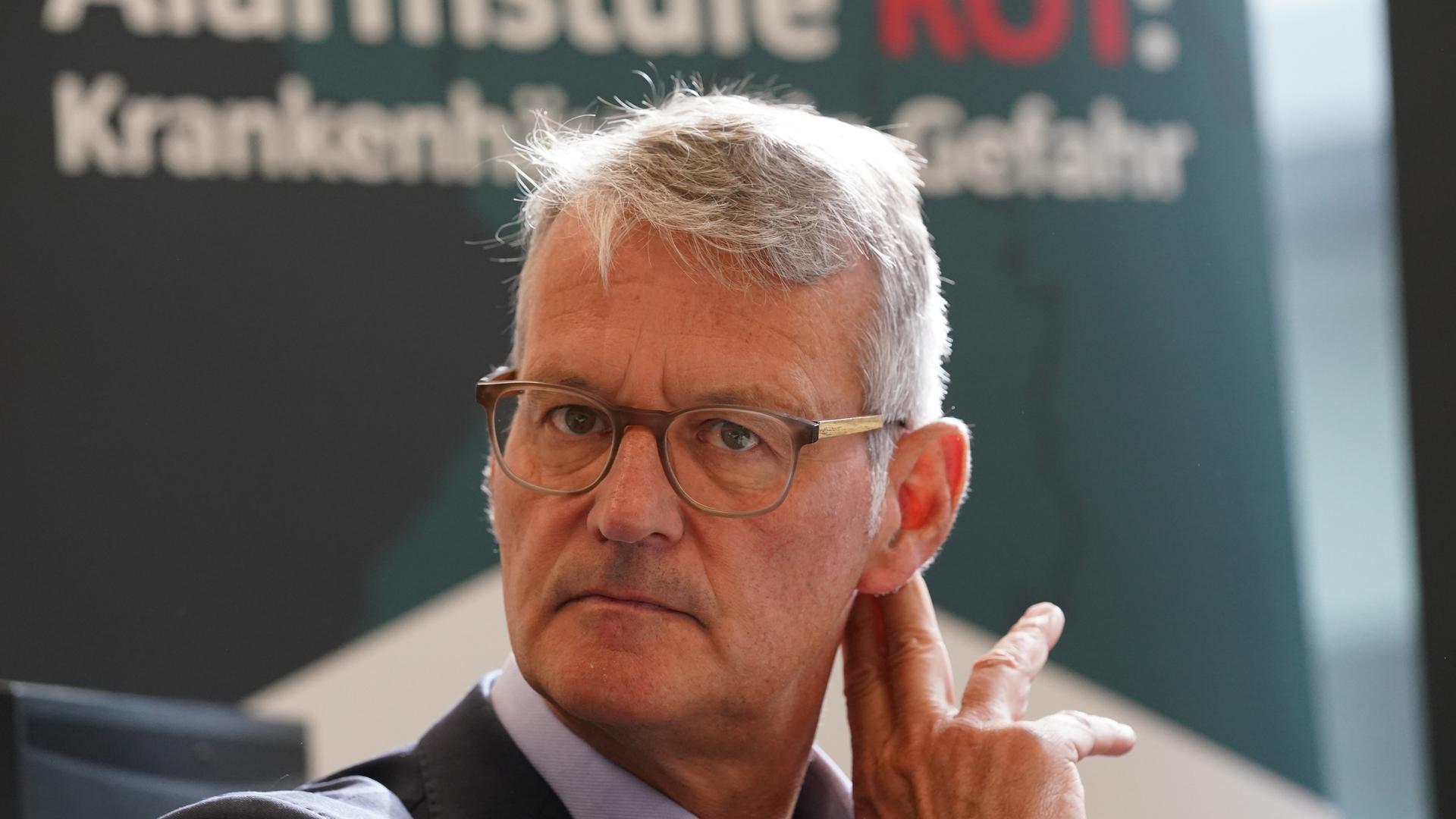 Gerald Gaß, Vorstandsvorsitzender der Deutschen Krankenhausgesellschaft (DKG), sitzt vor einem grünen Banner mit der weiß-roten Aufschrift: Alarmstufe Rot, Krankenhäuser in Gefahr
