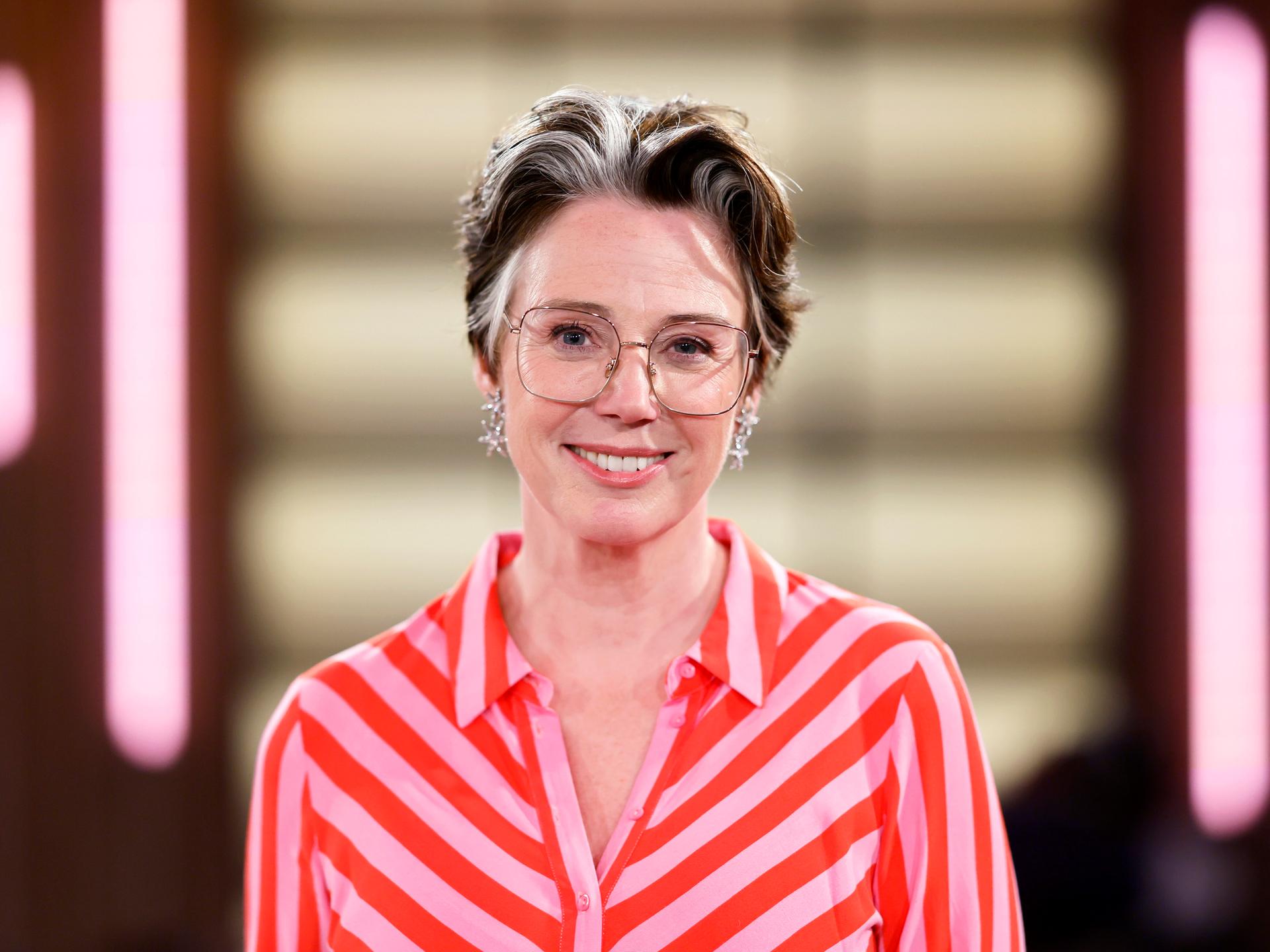 Ildiko von Kürthy steht in einem Fernsehstudio und lächelt in die Kamera. Sie trägt ein pink-orange-gestreiftes Kleid.