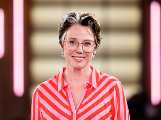 Ildiko von Kürthy steht in einem Fernsehstudio und lächelt in die Kamera. Sie trägt ein pink-orange-gestreiftes Kleid.