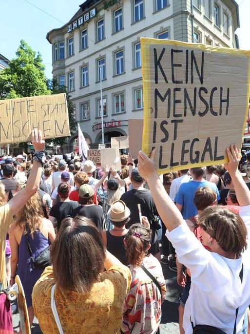 Menschen demonstrieren nahe einer Gedenkstele zum Gedenken an die Opfer einer Messerattacke von 2021 mit Plakaten auf denen "Menschenrechte statt rechte Menschen" und "Kein Mensch ist illegal" zu lesen ist, gegen eine Kundgebung der AfD.