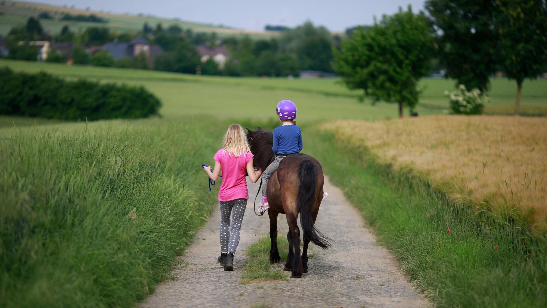 Ein Maedchen reitet unter Aufsicht mit einem Pony auf einem Feldweg aus.